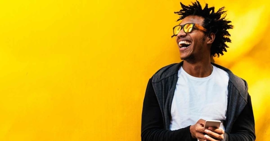 A man standing in front of a yellow wall. He is smiling and wearing sun glasses.