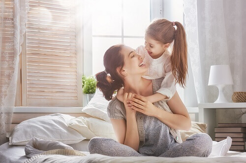 A mother with child custody sits on a white bed with her young daughter leaning over her shoulder. Both are smiling at each other.